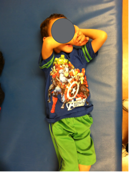 A young boy who is a hands dependent sitter lying supine on a therapy table with his forearms covering his face